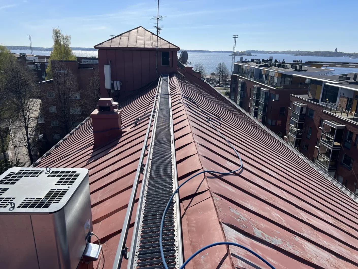 Hotellihoone kulunud punakaspruun valtsplekk-katus koos katuse käiguteega, meri ja linnasiluett taamal