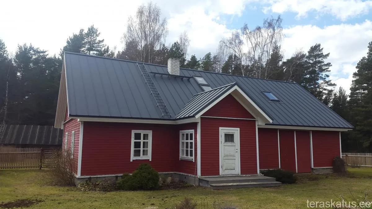 Punane talumaja tumeda valtsplekk-katuse, katuseredeli ja kahe katuseaknaga
